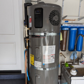 Gray water heater in a garage beside blue water filters and a white door.