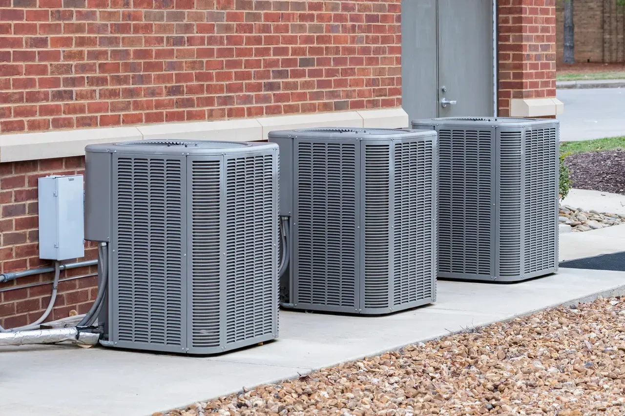 Three gray outdoor HVAC units beside a brick building on a concrete pad