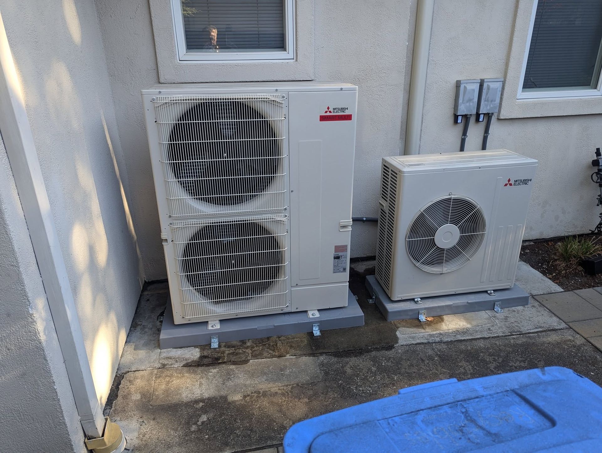 Two Mitsubishi mini-split outdoor units beside a wall, with a blue bin in the foreground.