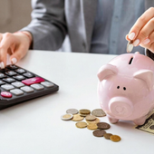 Person counting coins beside a pink piggy bank and calculator on a desk