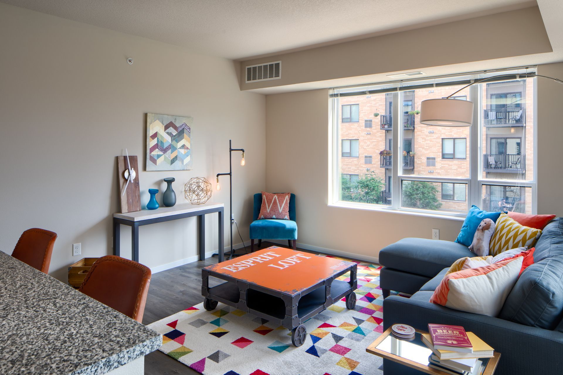 Modern living room with blue sofa, colorful rug, and a large window overlooking buildings.