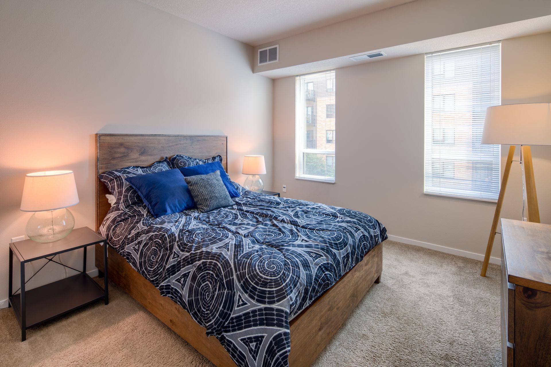 Bedroom with a bed, nightstand, lamps, two windows with blinds, and a floor lamp.