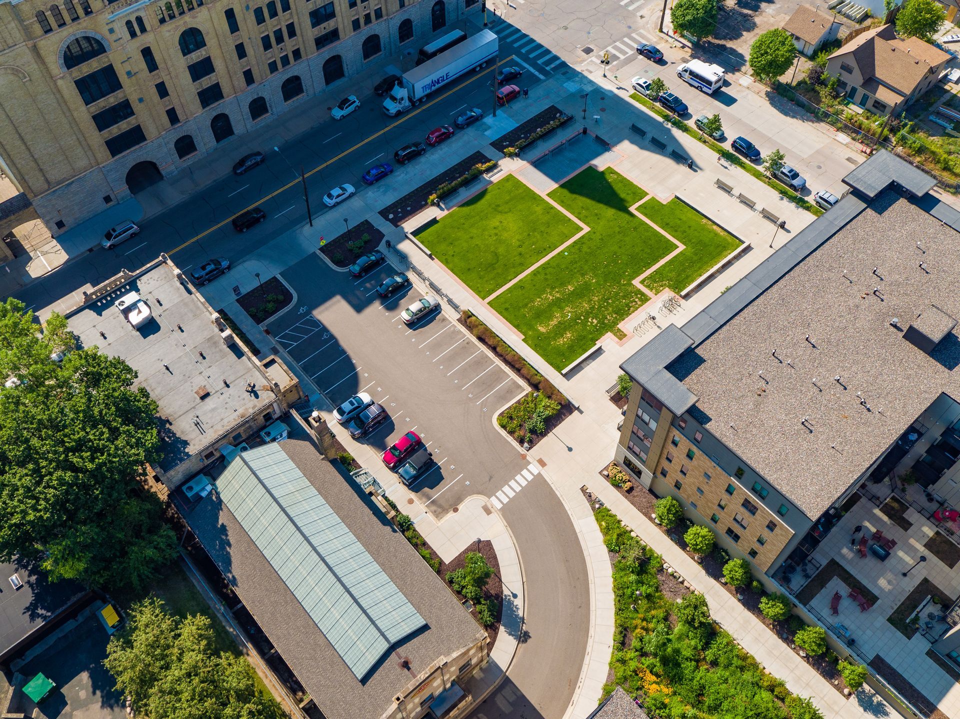 Aerial view of a city square with buildings, a green lawn, parking, and a crosswalk. Sunny day.