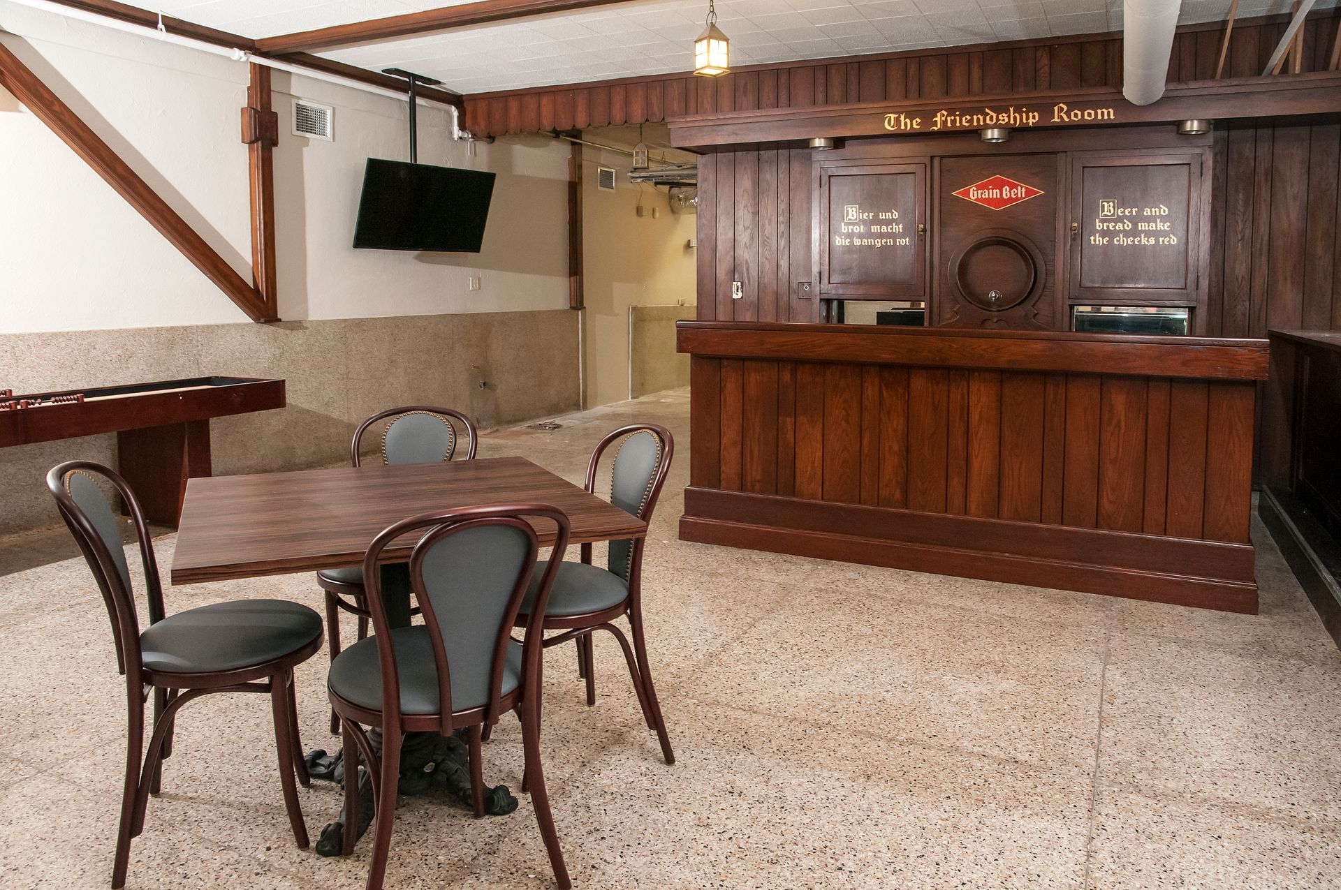 Bar interior with wooden bar, table with chairs, TV, and foosball table on a speckled floor.