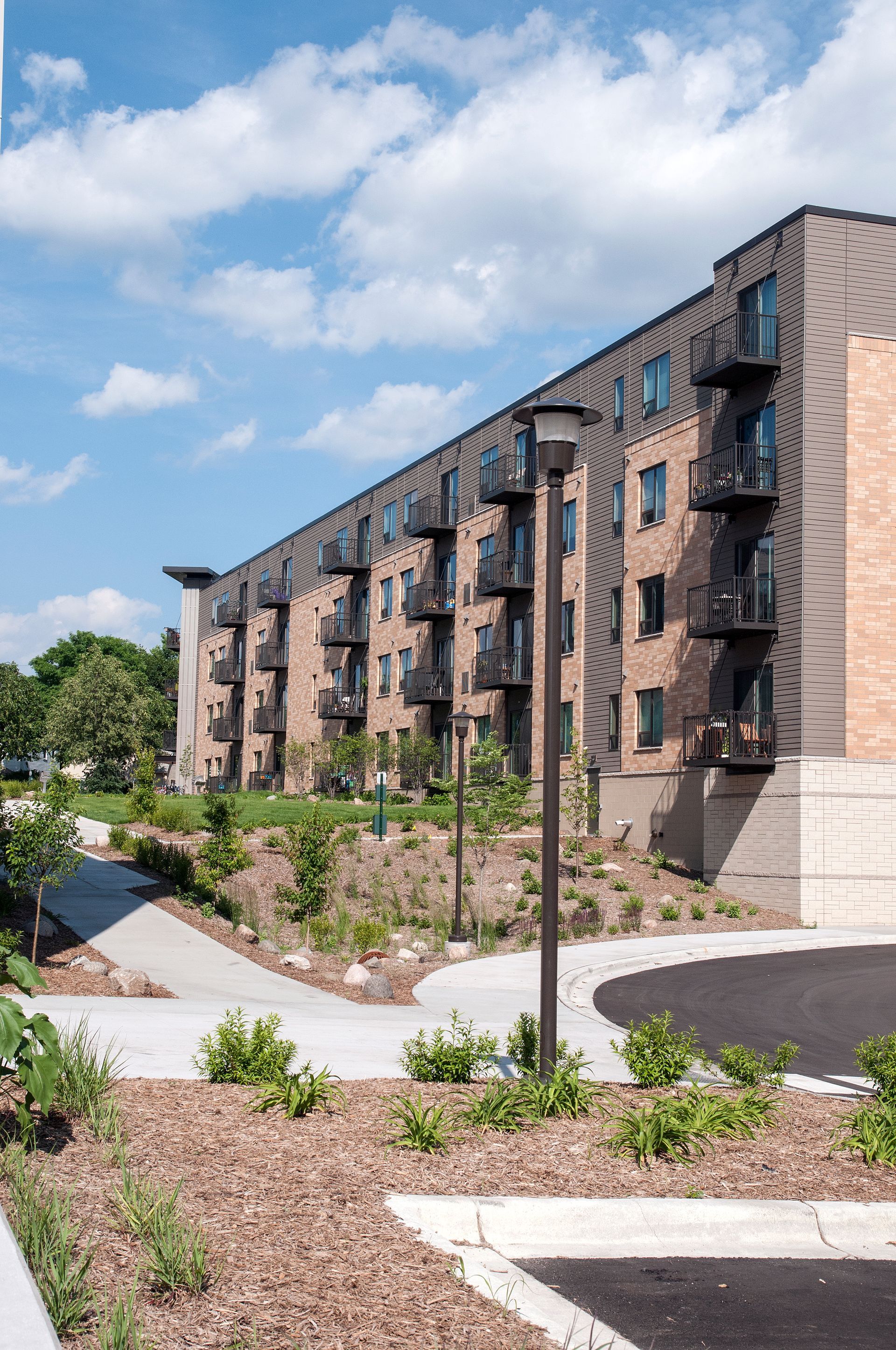 Multi-story brick apartment building with balconies, landscaped grounds, and a cloudy blue sky.