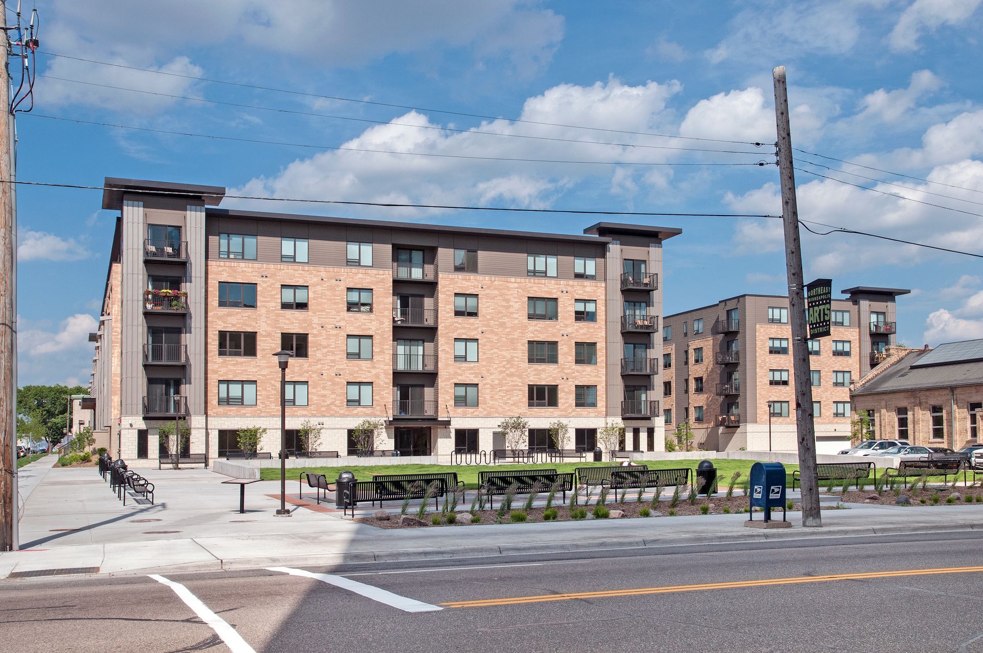 Multi-story brick apartment buildings with balconies, blue sky, and a small park.