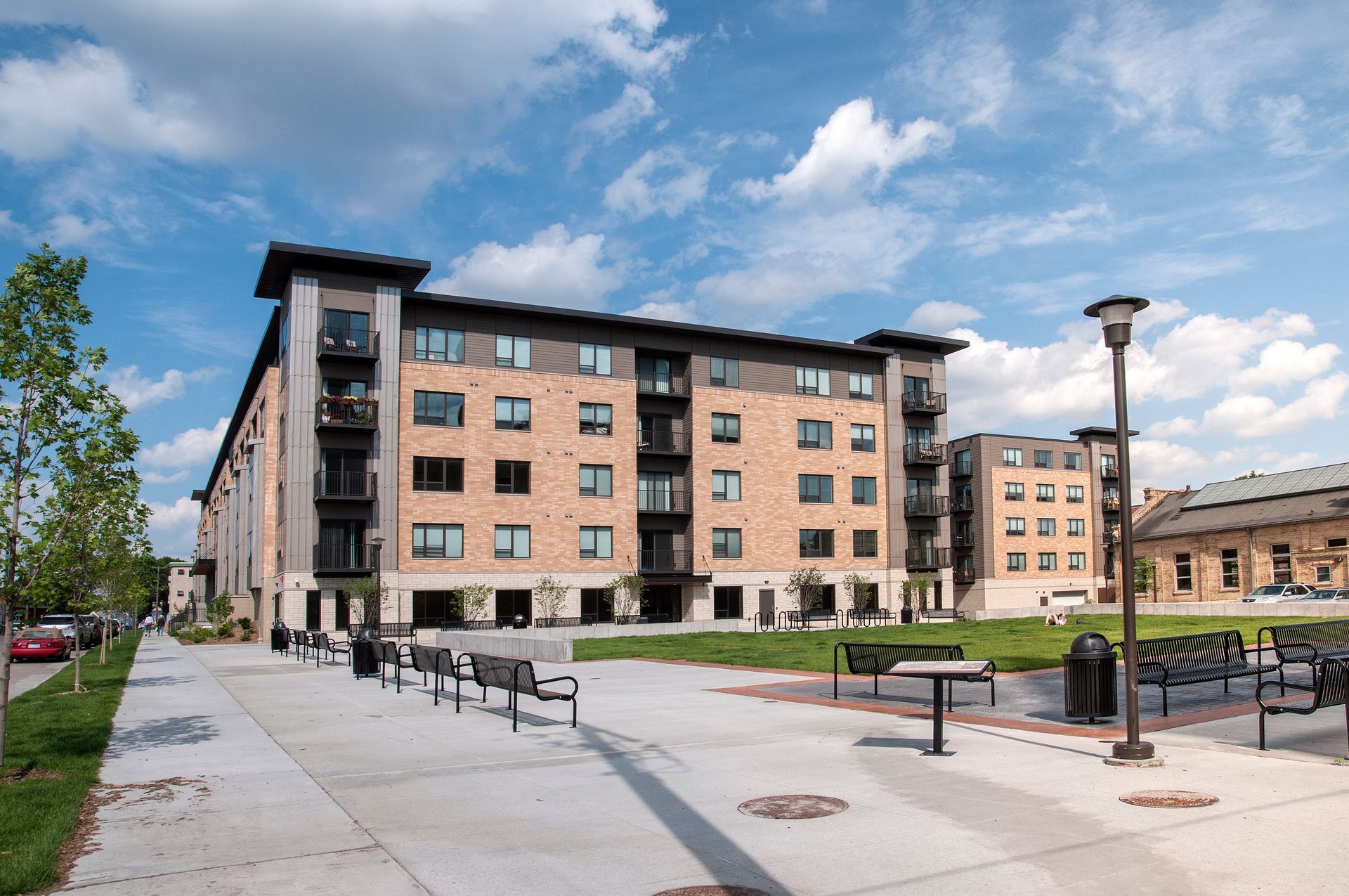 Multi-story brick apartment building on a sunny day with benches and a sidewalk in front.