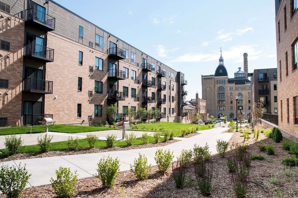Brick apartment buildings with balconies, pathway, and historic building in the background on a sunny day.