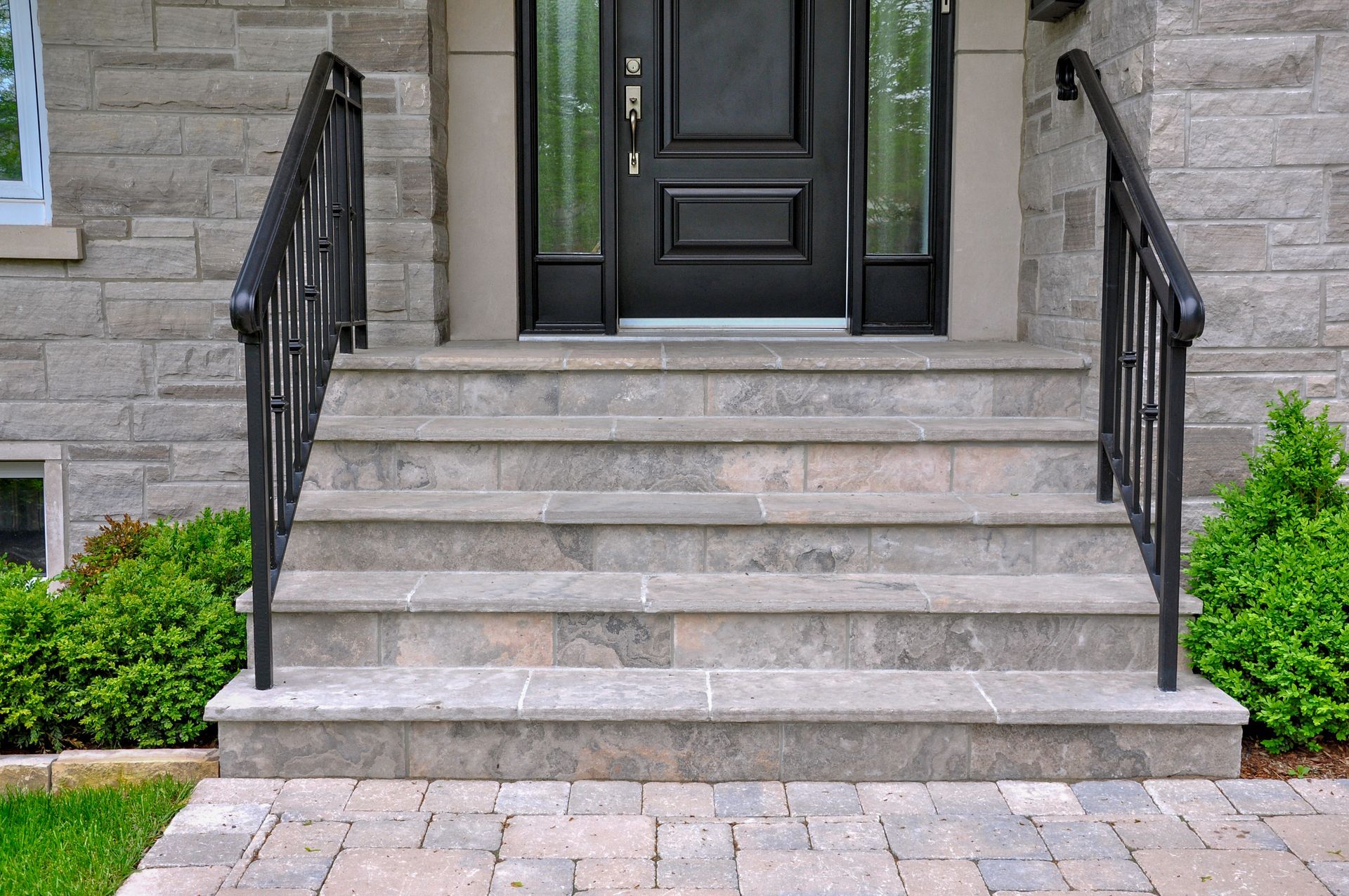 Stone front steps leading to a black front door with wrought-iron railings and trimmed shrubs on either side