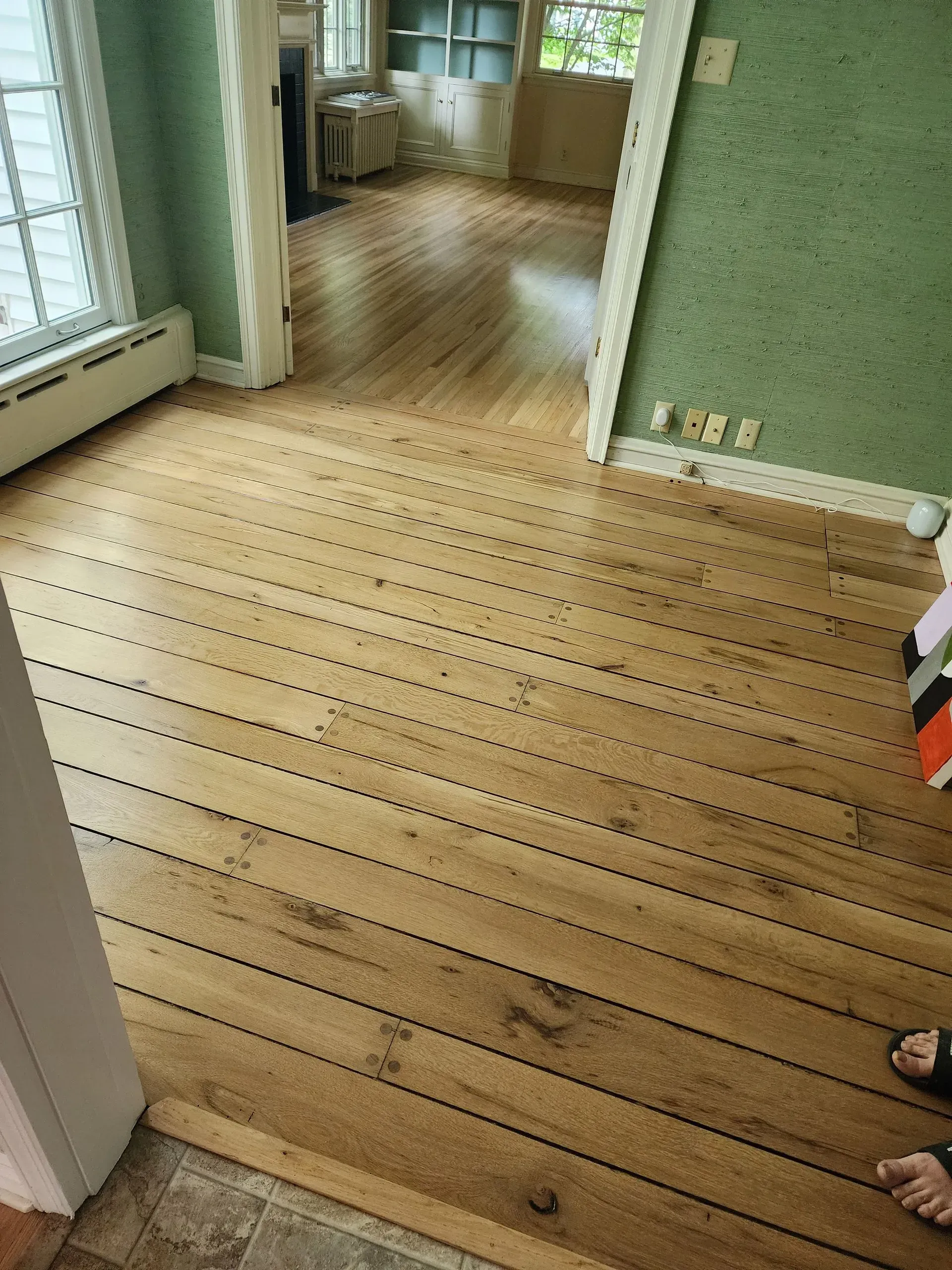 Wooden floor in a green room with a doorway and partial view of a person’s feet at right.