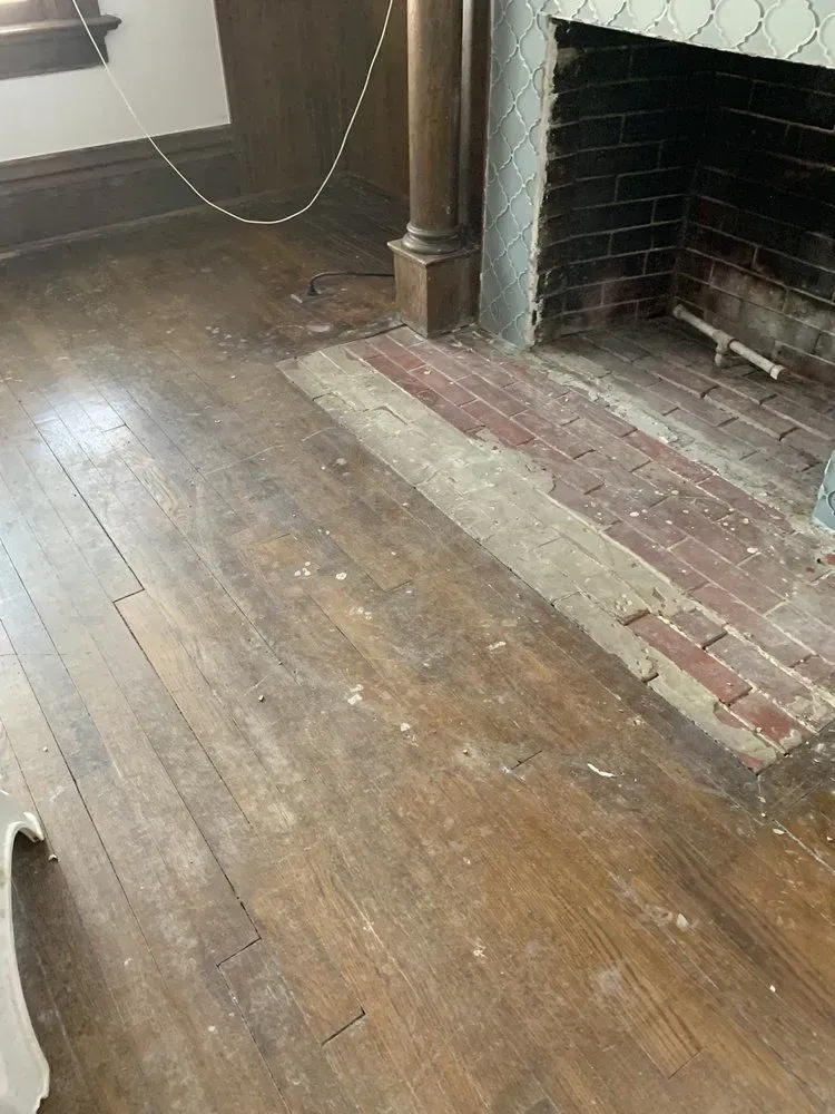 Old wooden floor beside a brick fireplace hearth in a dim room