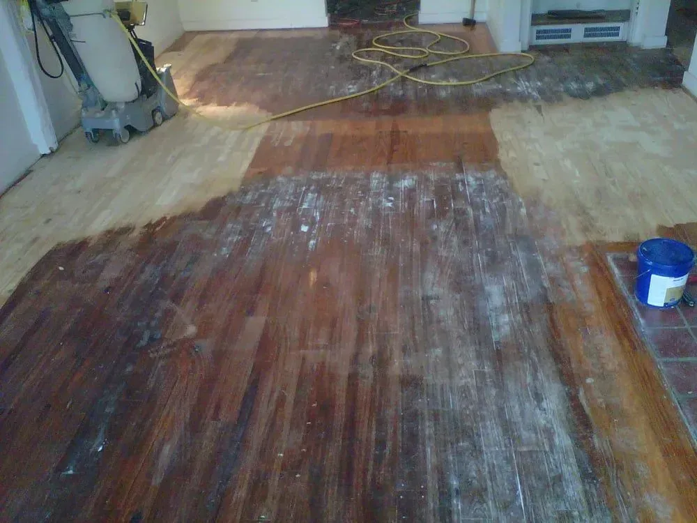 Partially sanded hardwood floor with white dust, extension cords, and a blue paint bucket in a room