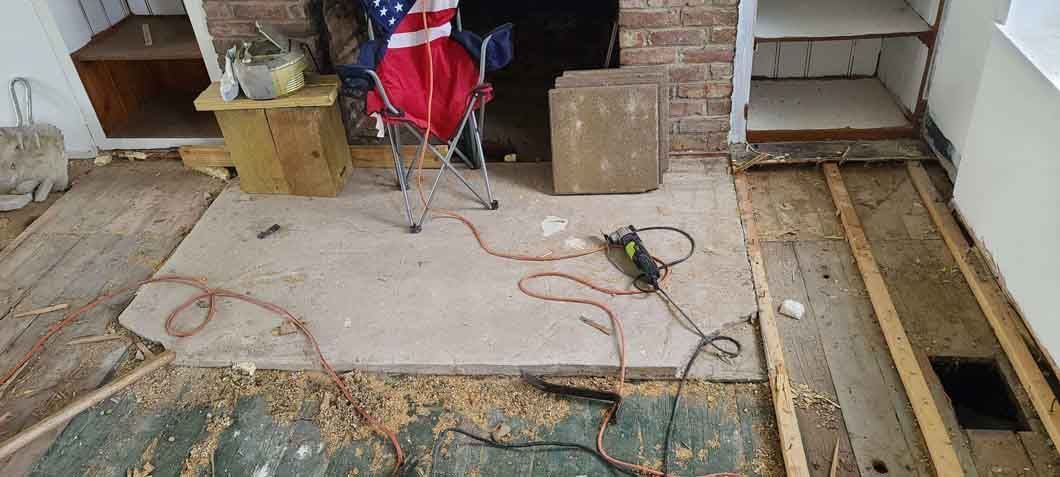 Partially renovated room with exposed subfloor, tools, cords, and a red folding chair near a fireplace