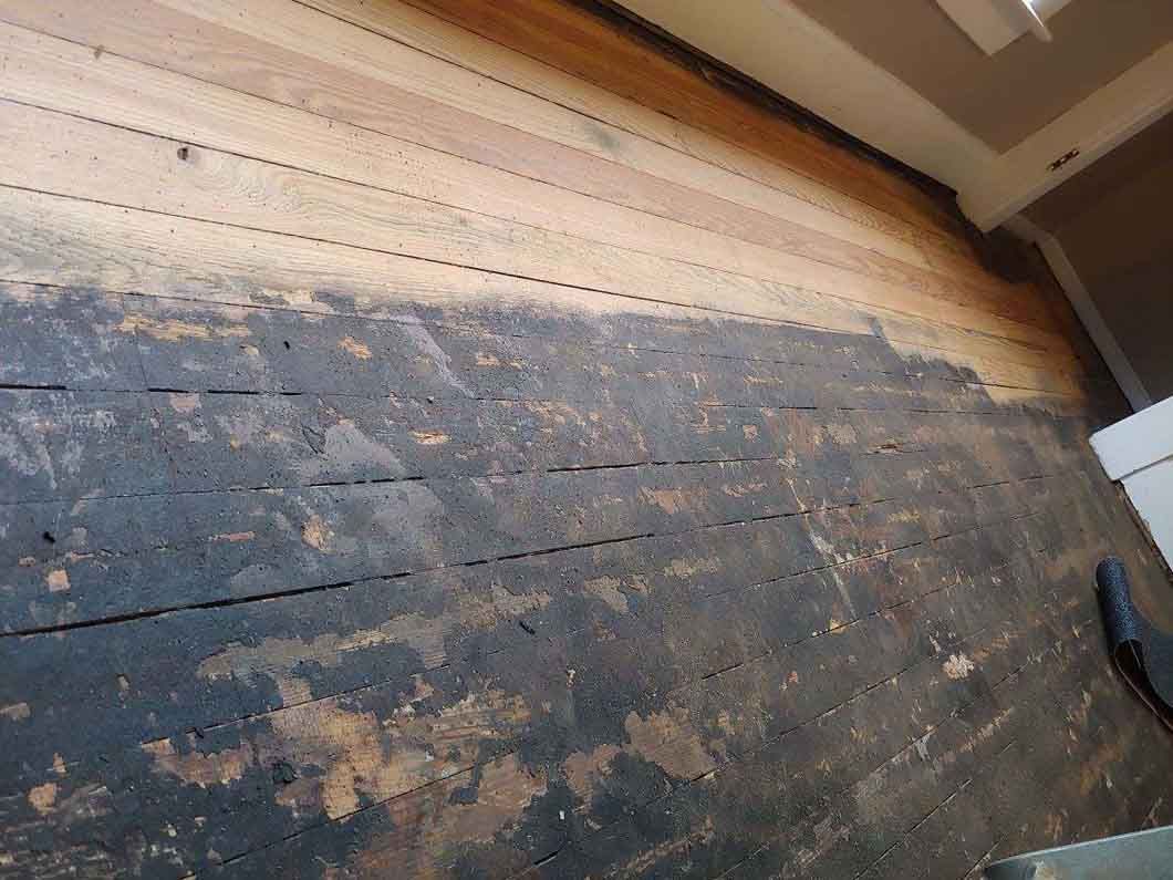 Worn wooden floorboards with dark stains and chipped paint near a wall and window trim