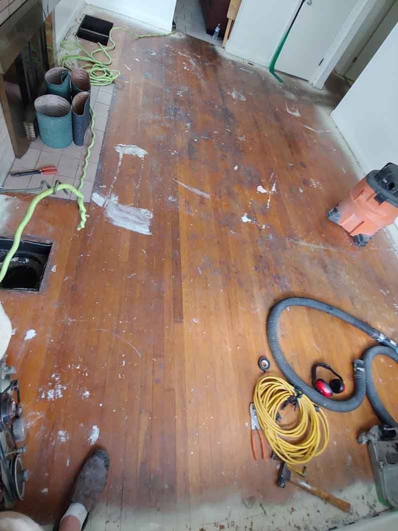 Wooden floor in a room under renovation, with tools, cords, and paint patches scattered around.