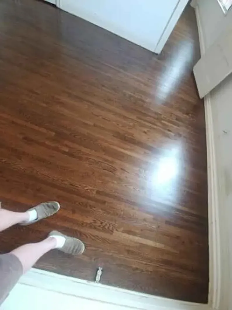 Wooden floor in a room, with a person’s feet in gray shoes at the bottom left.
