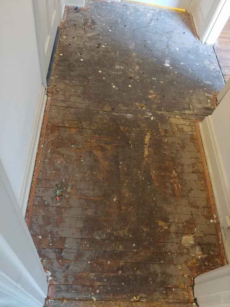 A narrow hallway with a worn, debris-covered floor and white walls, lit from a doorway at the end.