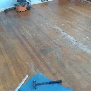 Wooden floor with water damage, a mop and hammer on the floor near the wall.