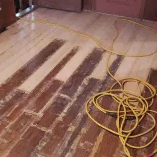 Wooden floor being sanded with dark worn strips and a yellow extension cord coiled on the right