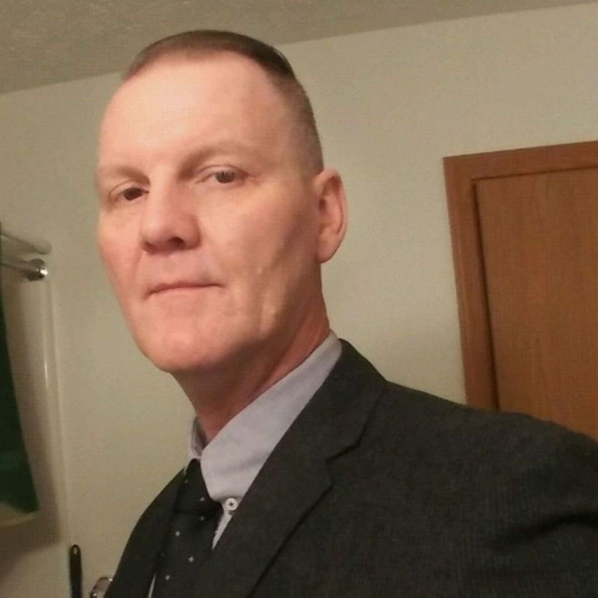 Man in a dark suit and tie posing indoors beside a wooden door