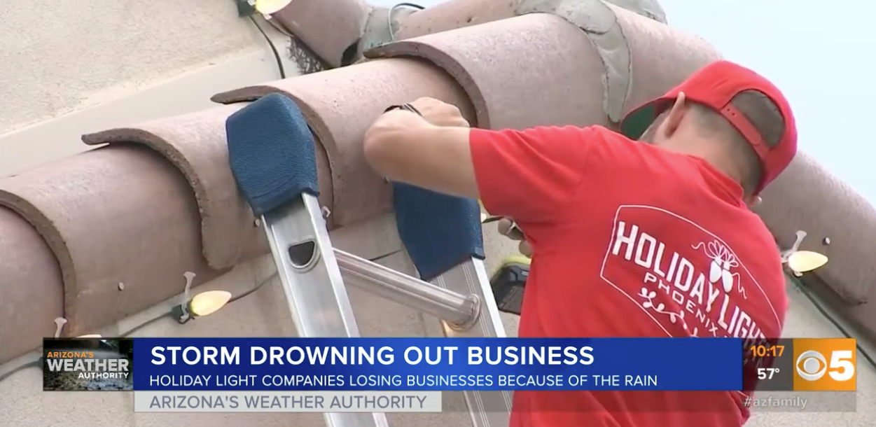 Worker in red shirt inspects storm drain on a bridge as news banner reads “Storm drowning out business”