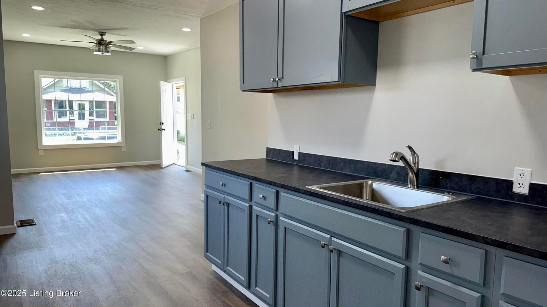 Empty kitchen with gray cabinets, black countertop, and open view to a bright living area with ceiling fan.