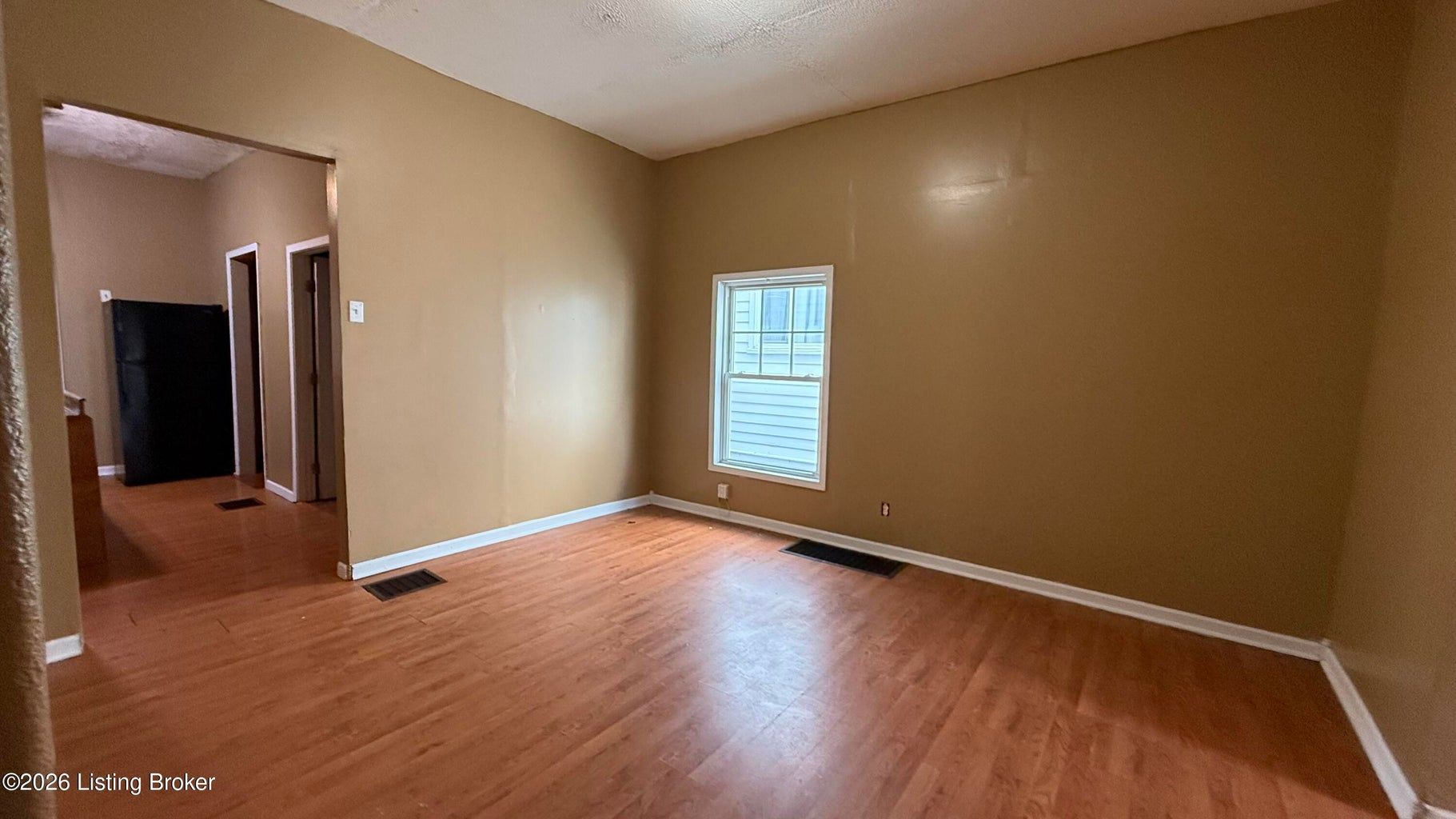 Empty beige room with wood floors, one window, and a doorway opening to a hall or closet