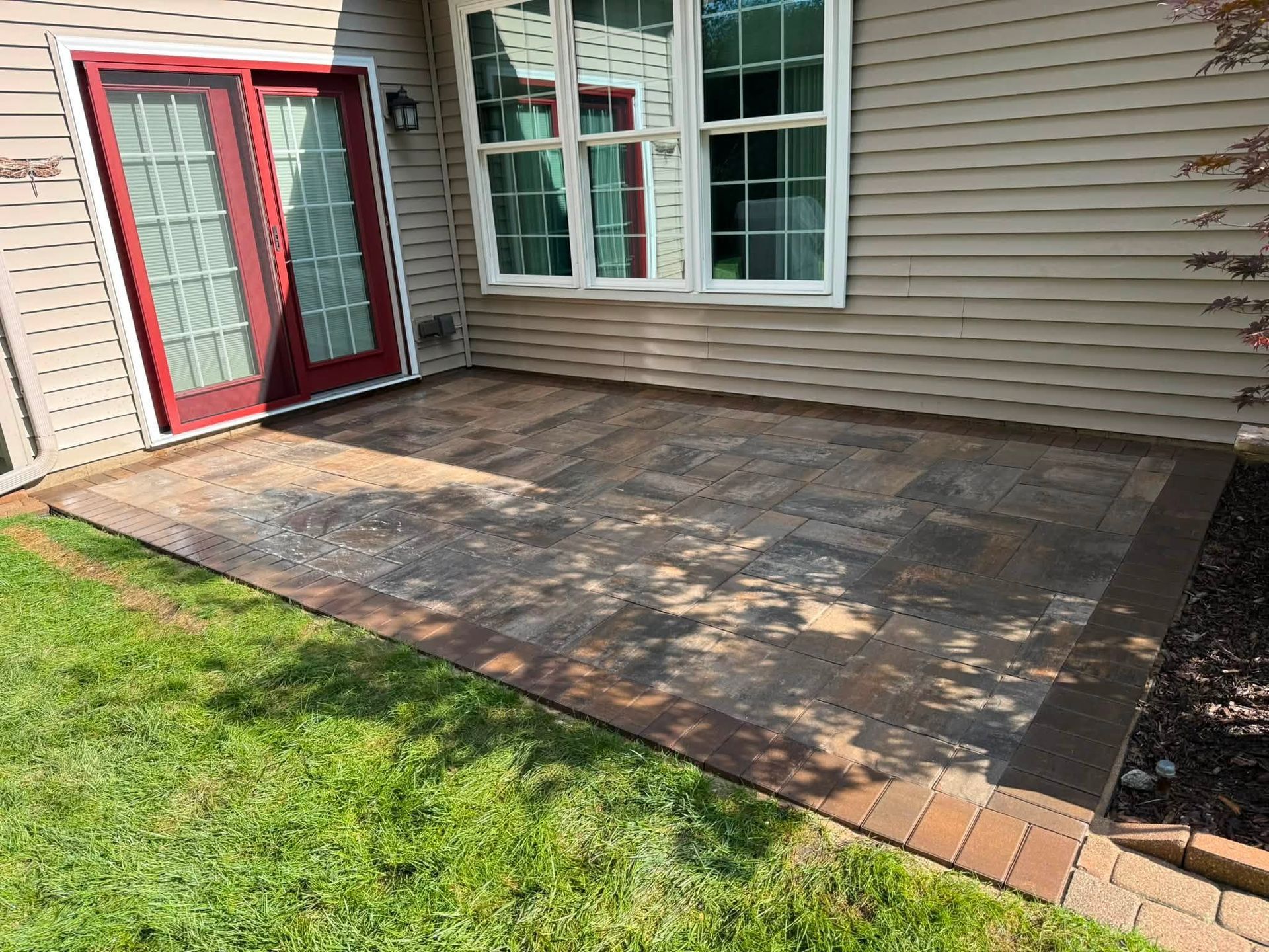 Backyard patio with faded stone surface beside beige house and red double doors