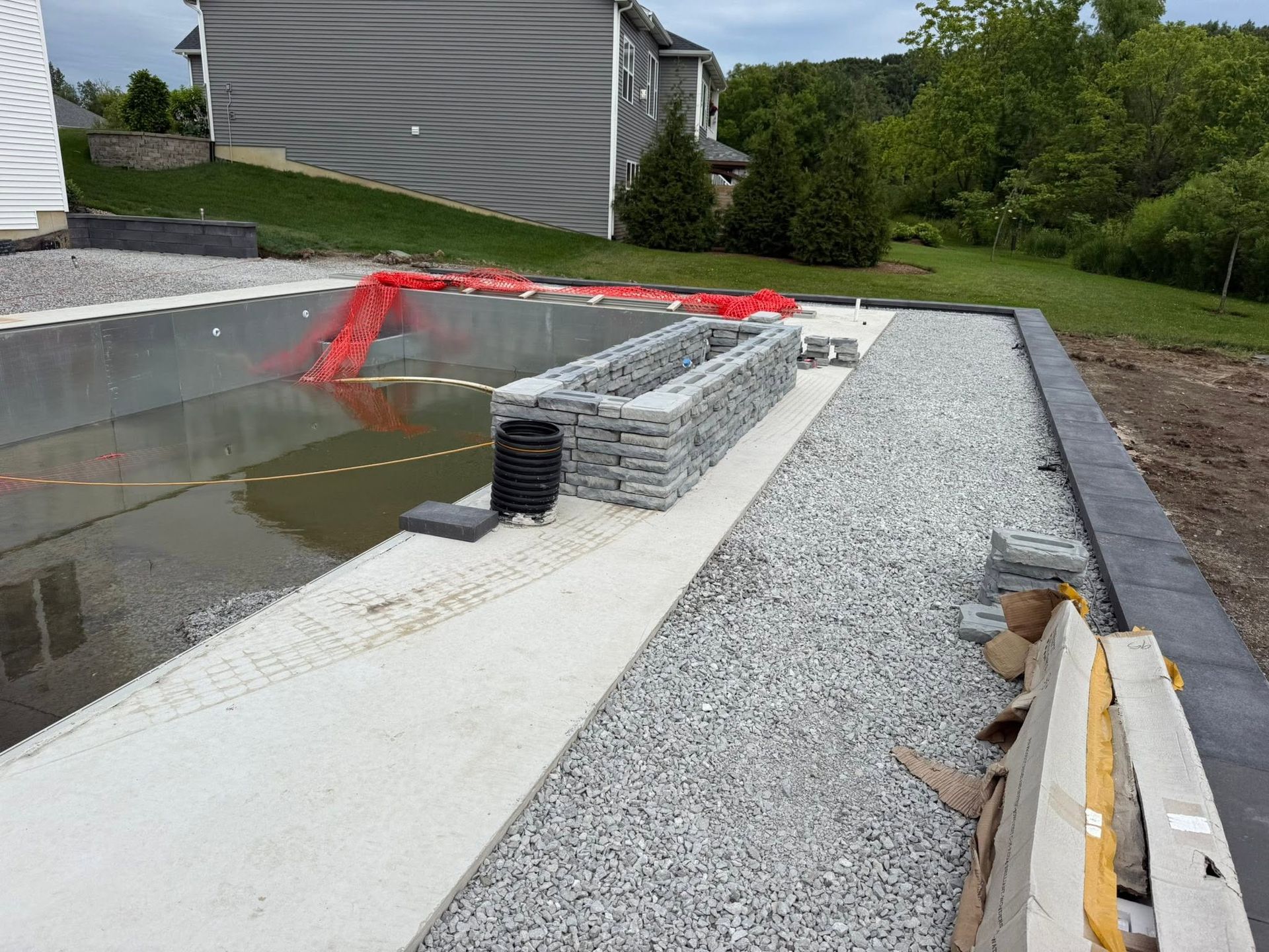 Backyard construction site with gravel trench, concrete slab, and stacked pavers beside a house