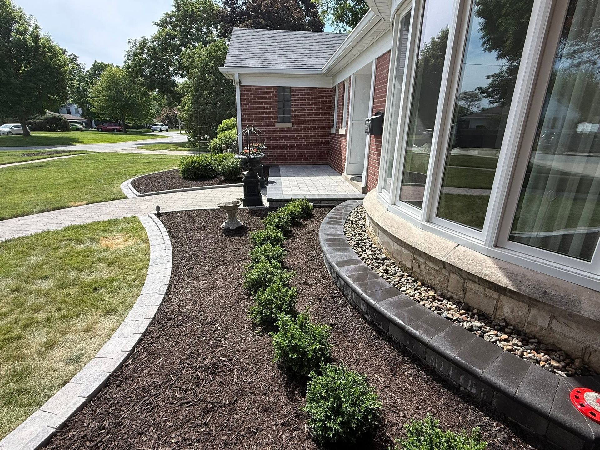 Curved garden bed with dark mulch and small shrubs beside a brick house and large windows