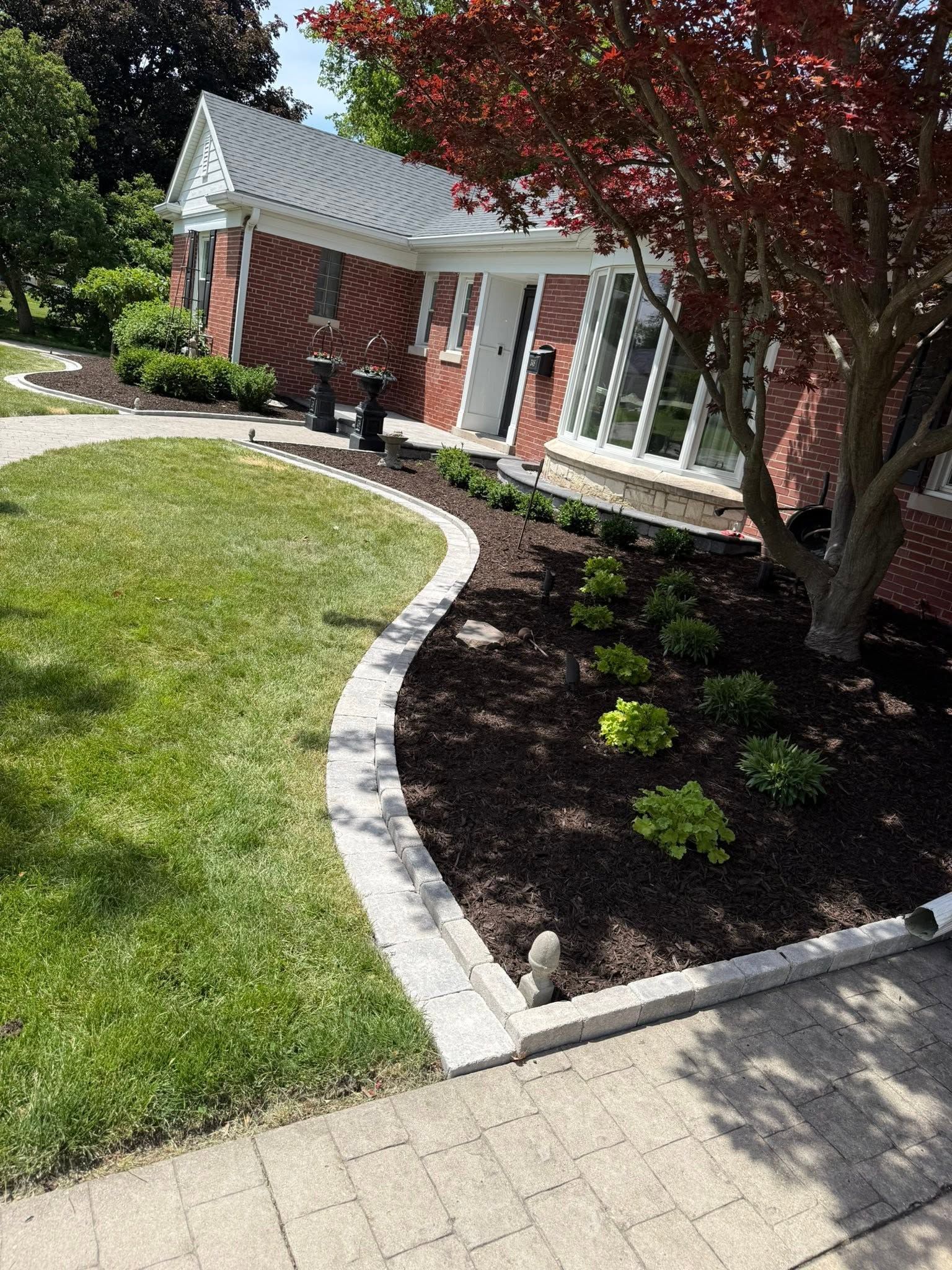 Curved stone border around a green lawn and mulched flower bed beside a red brick house and tree