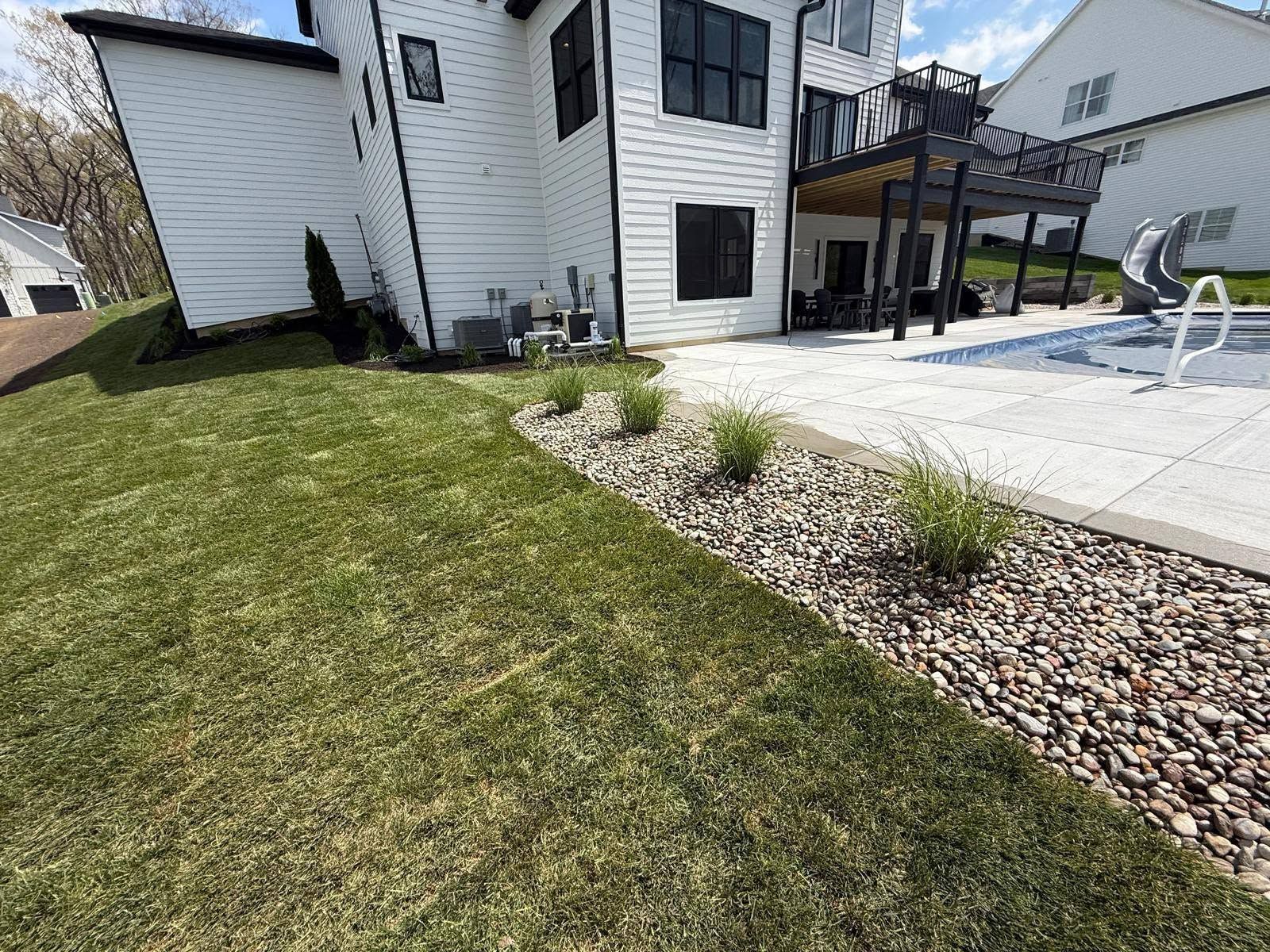 Backyard with green lawn, white house, stone border, and patio beside a small pool