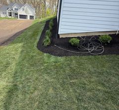 Side yard with a black mulch bed, coiled garden hose, and white house beside a driveway.