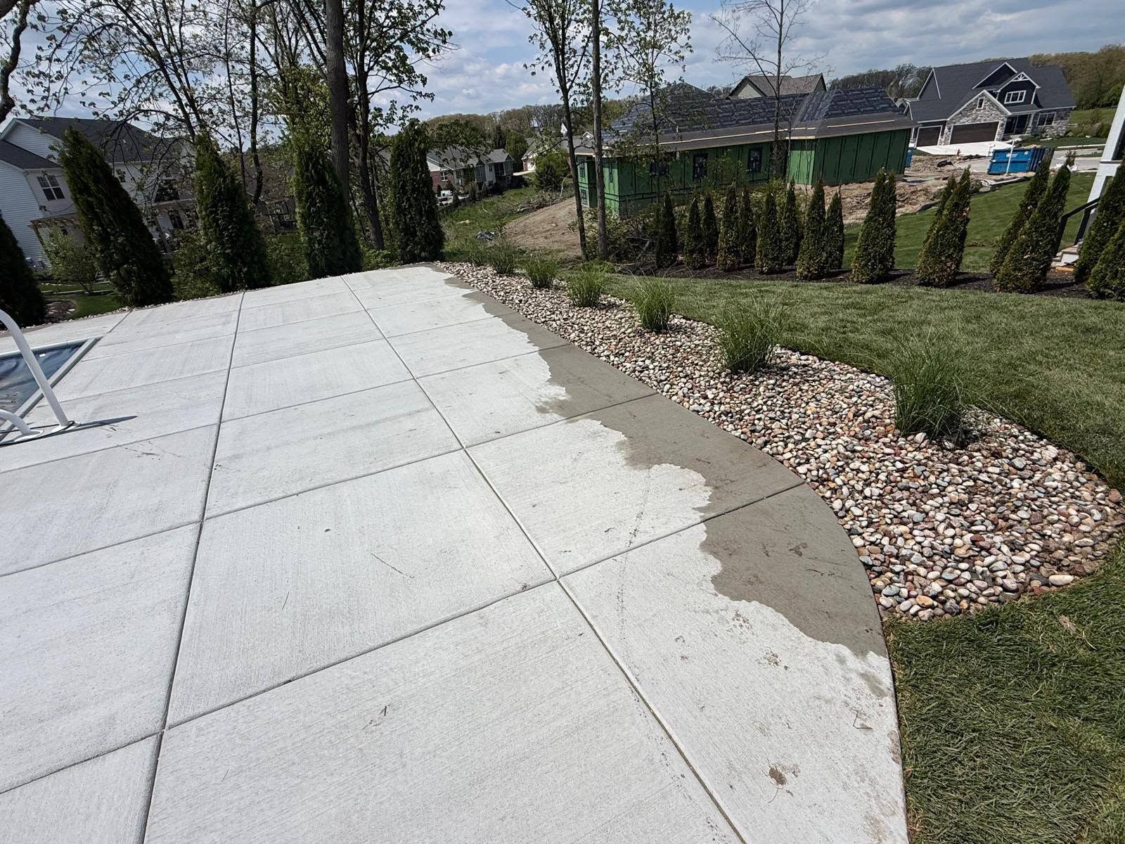 Curved concrete patio beside a landscaped yard with rocks, grass, and trees on a sunny day