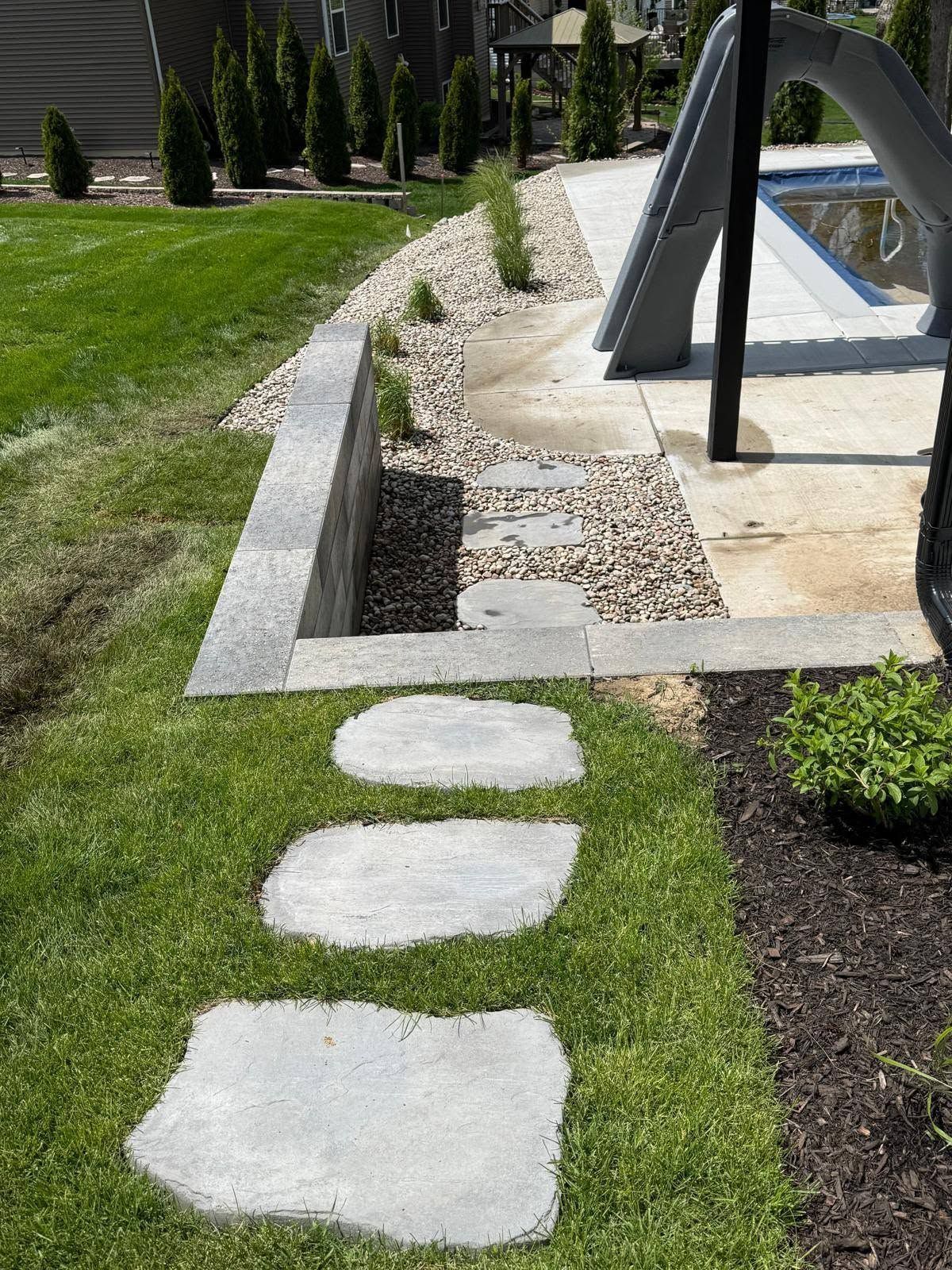 Stepping stones in a green lawn leading to a gravel path beside a modern poolside structure.