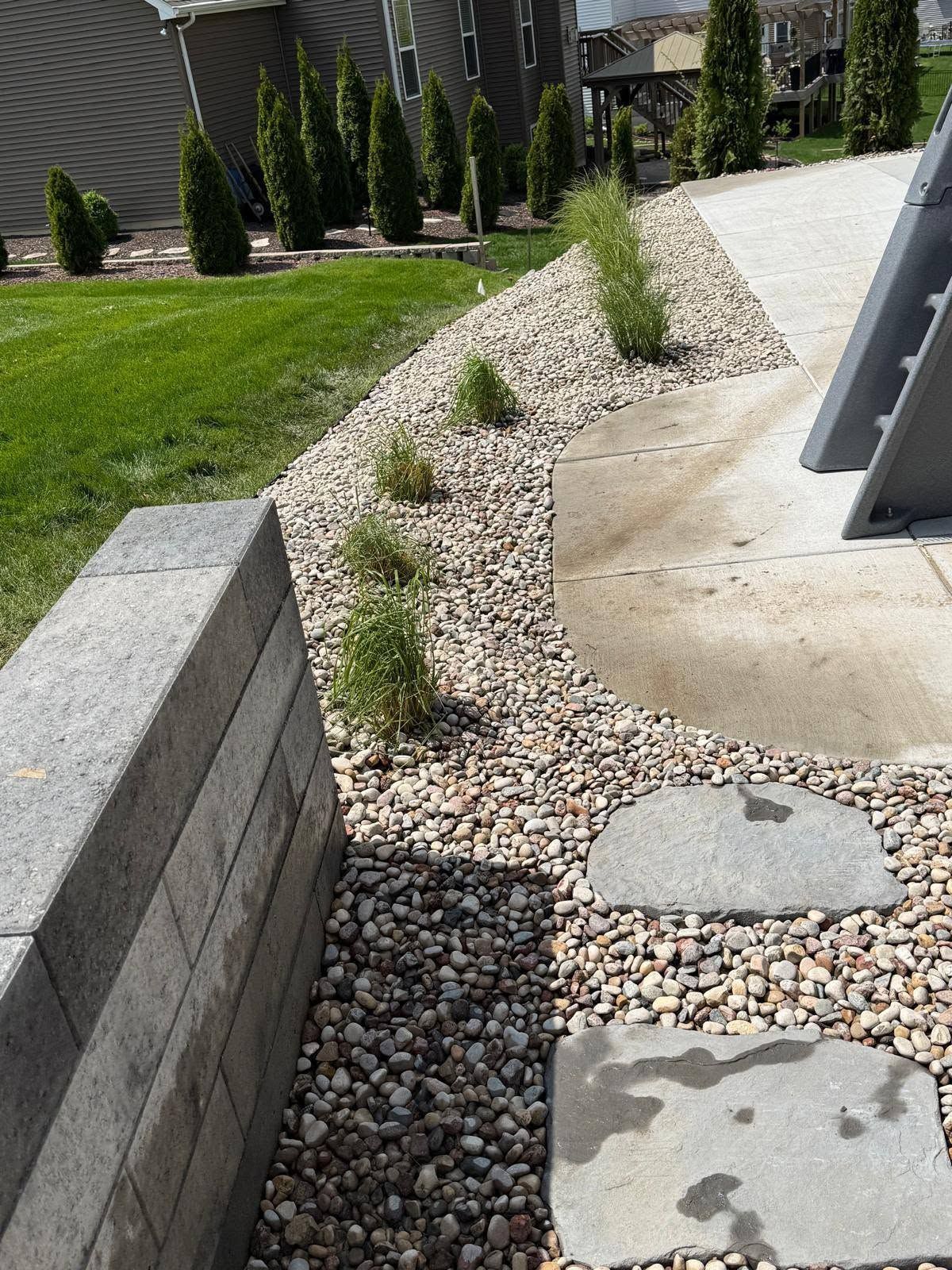Landscaped walkway with gravel, concrete pavers, and small shrubs beside a lawn and retaining wall