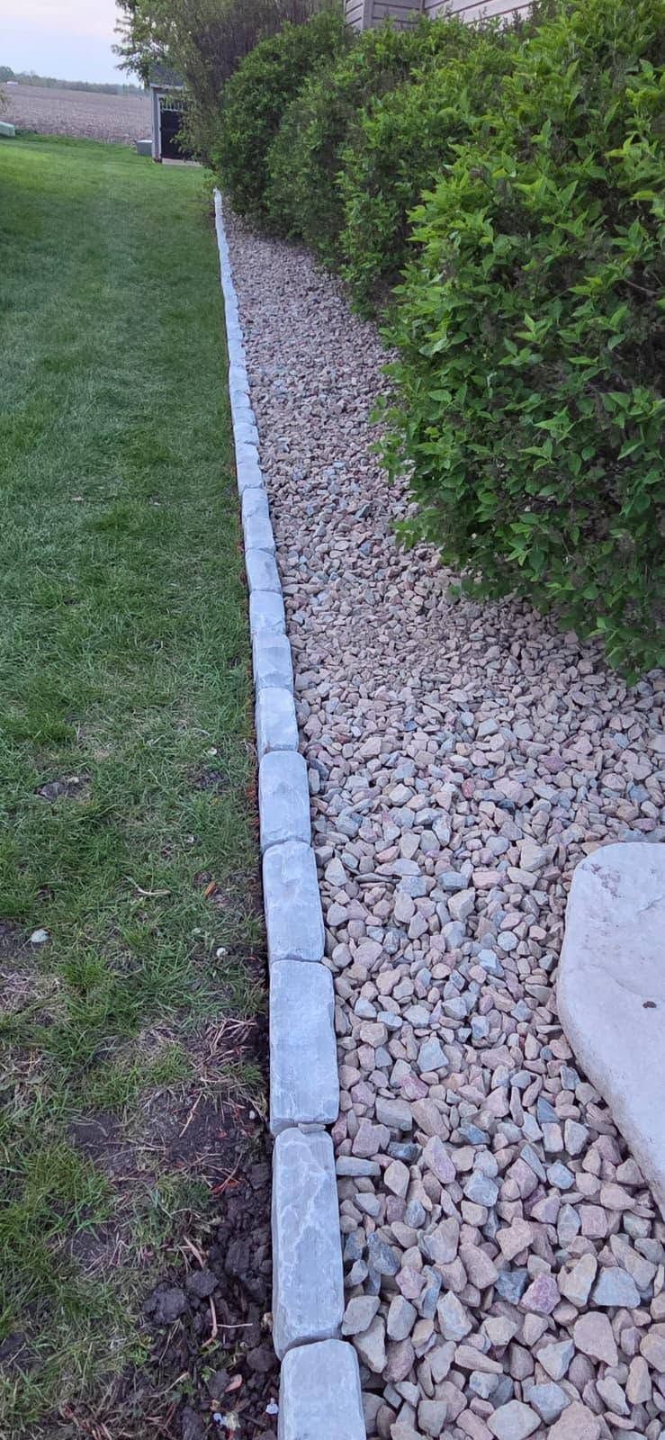 Narrow gravel border between green lawn and tall hedge, edged with concrete curb
