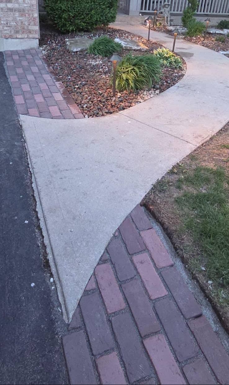 Curved concrete sidewalk bordered by brick pavers and a small landscaped bed with shrubs and mulch.