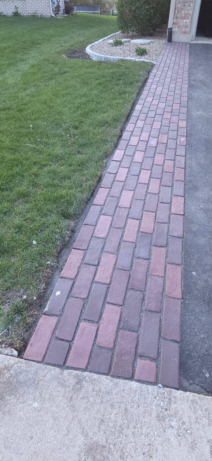 Brick walkway beside a green lawn leading to a house entrance, with a small landscaped bed.