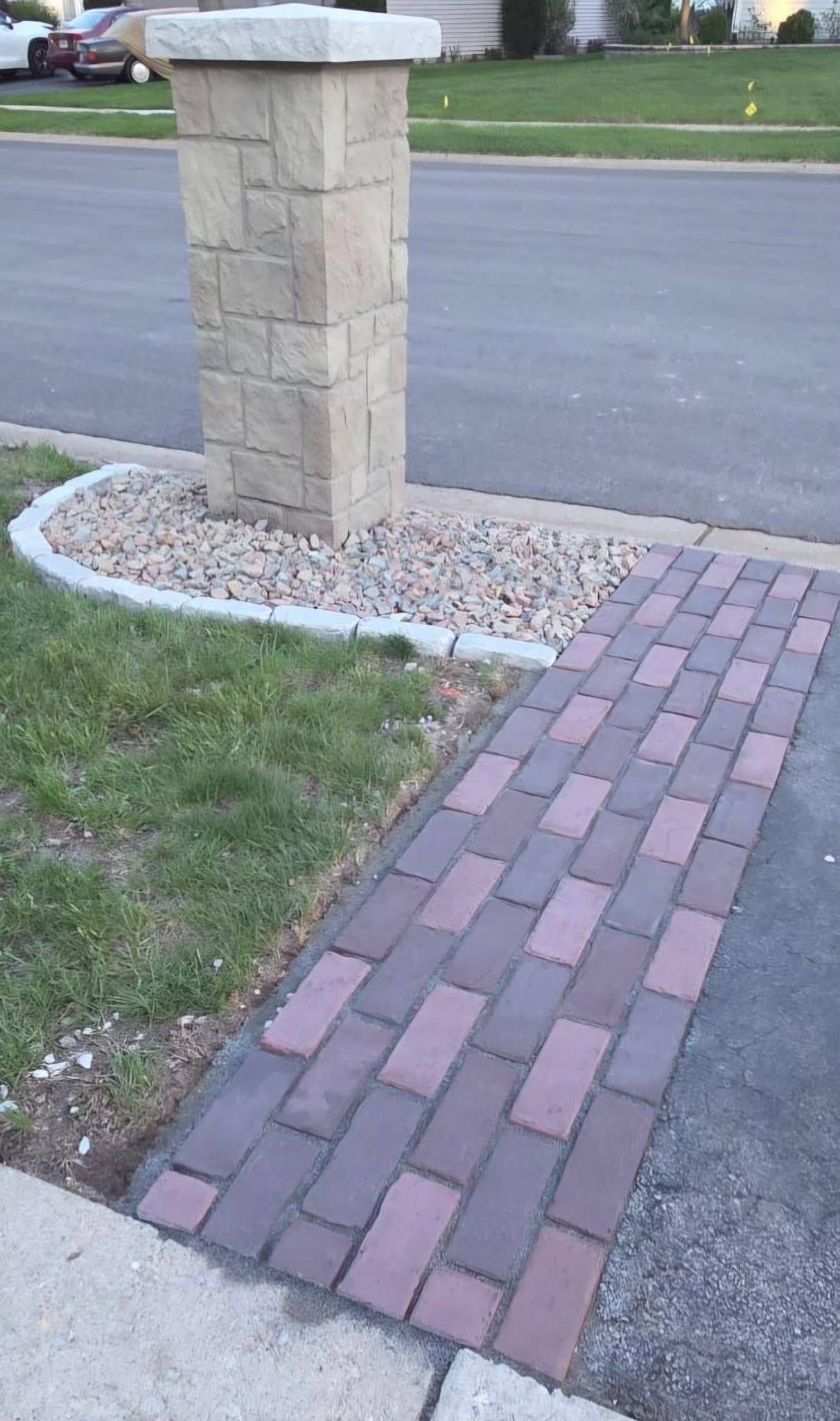 Brick sidewalk leading to a stone mailbox pillar beside a street and gravel bed