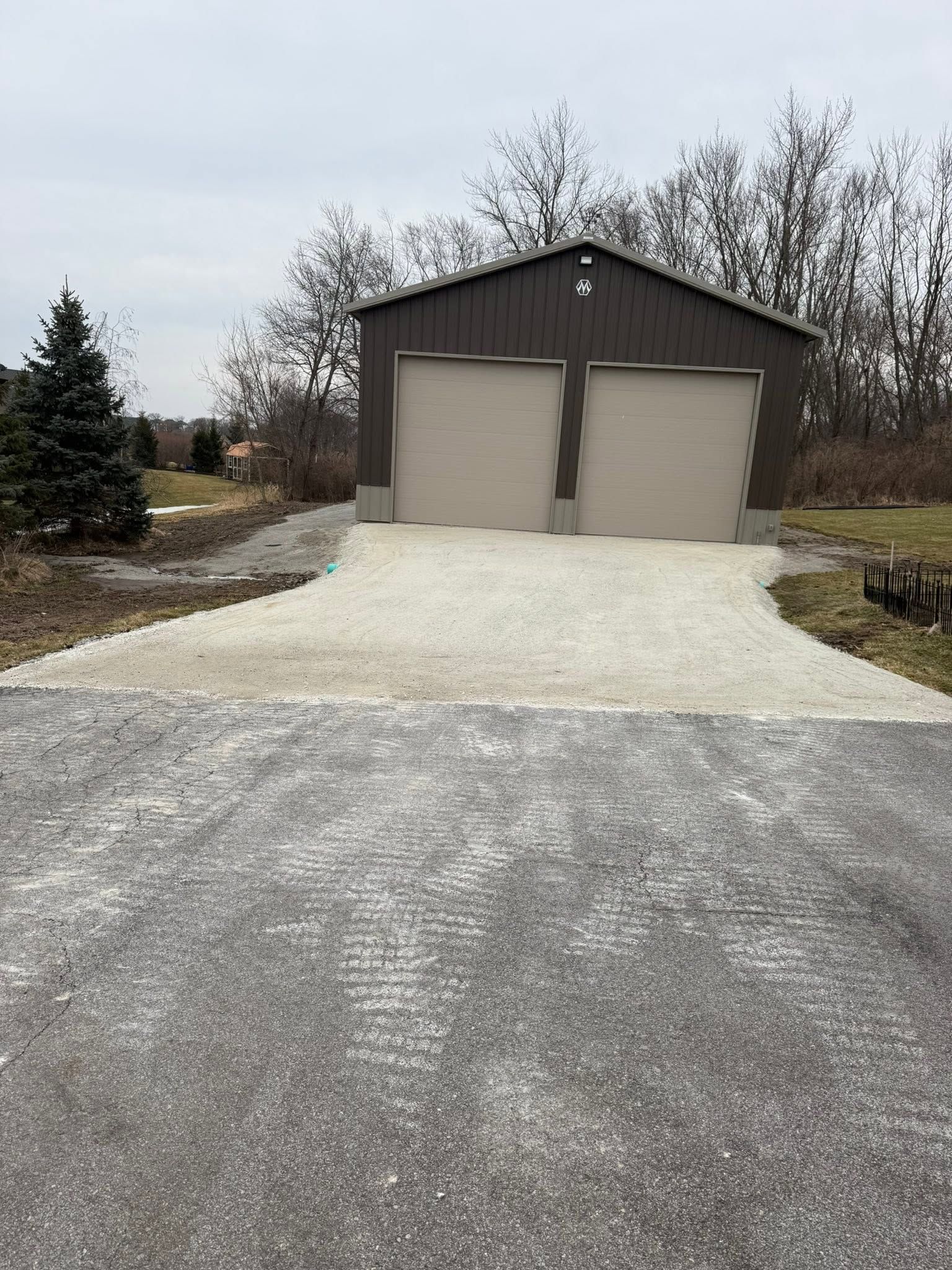 Gravel driveway leading to a brown garage with two closed doors and bare trees in the background