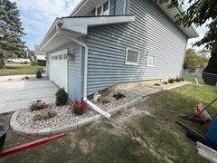 White house exterior with attached garage, gray siding, and a landscaped stone border along the foundation