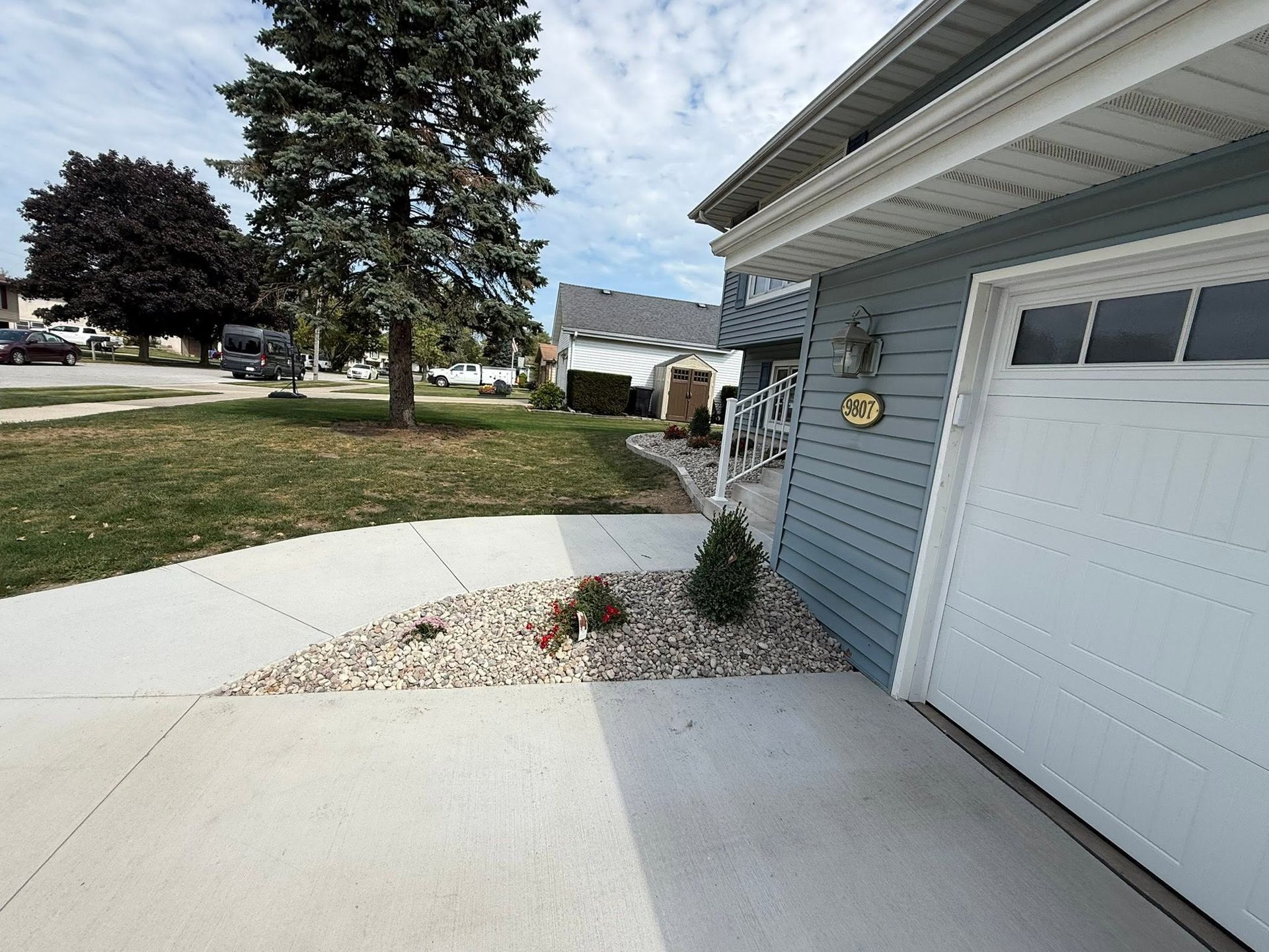 Driveway beside a gray house with white garage door and a curved concrete walkway edged with rocks and shrubs.