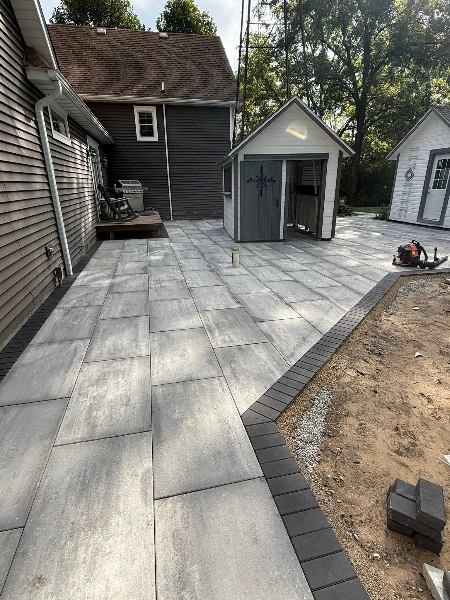 Backyard patio with large gray pavers, a small shed, and a dirt garden bed under construction