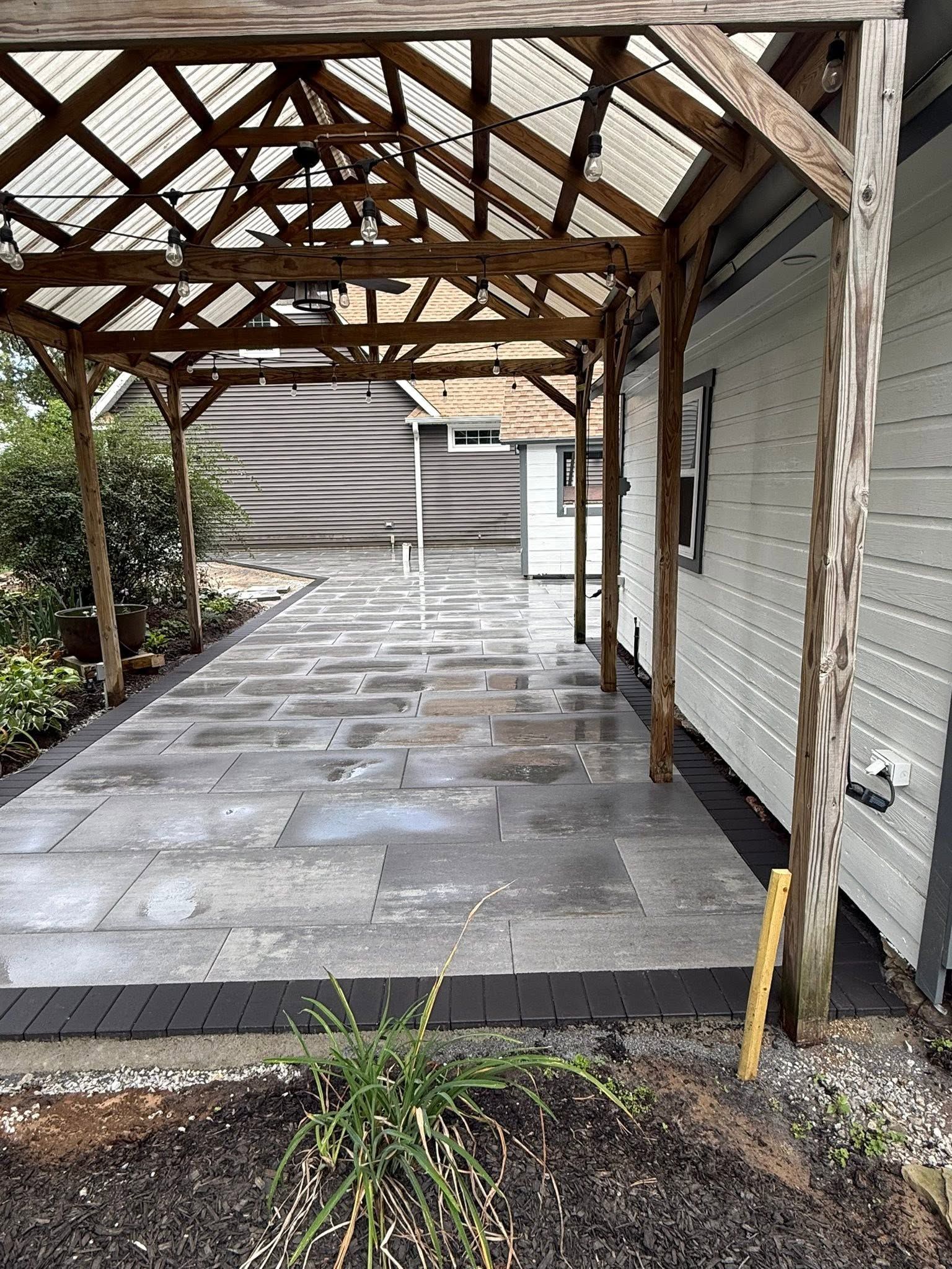 Covered stone patio with a wooden pergola, damaged roof beams, and a white house wall.