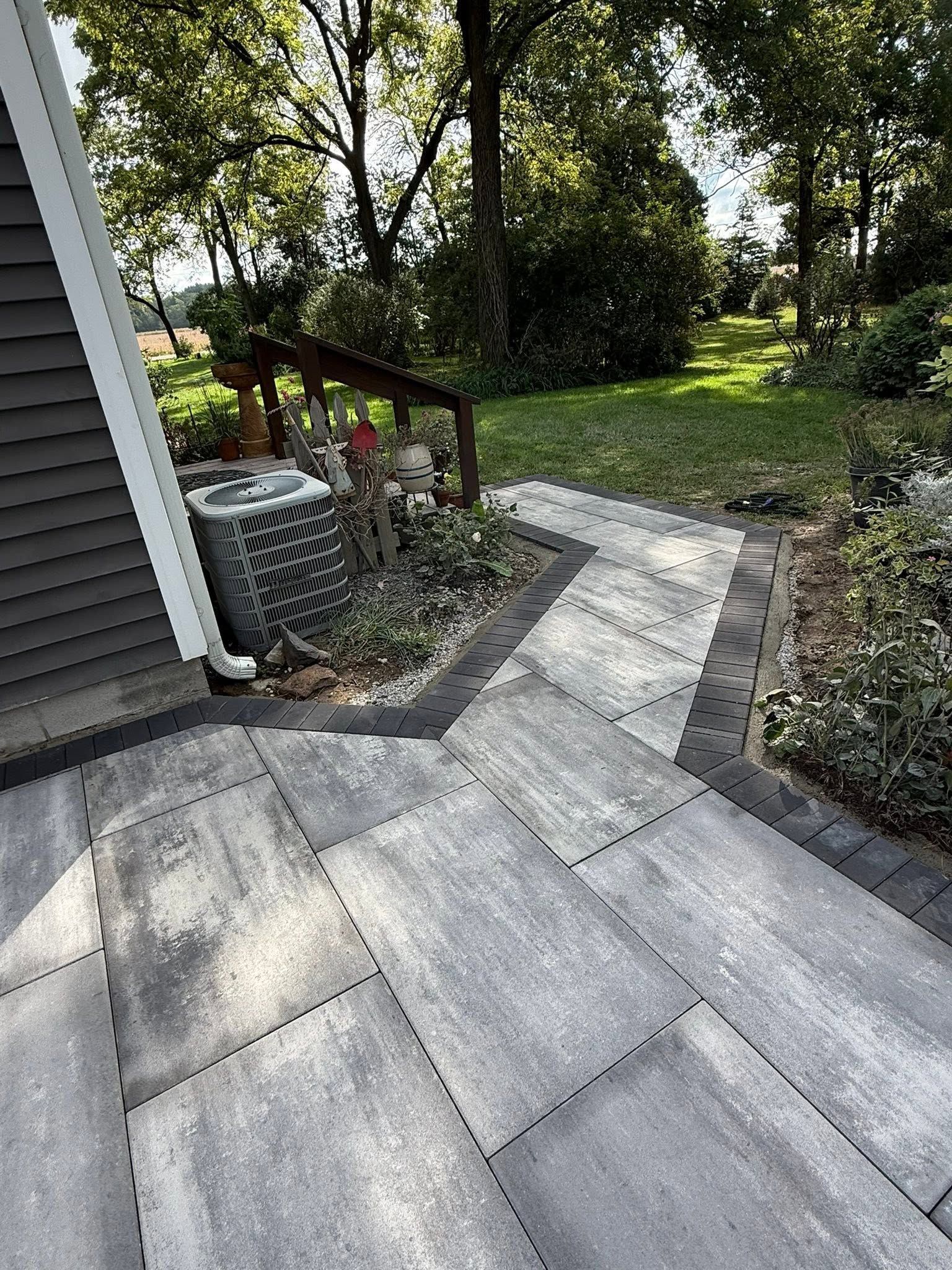 Stone patio walkway beside a house leading into a green backyard with trees and shrubs.