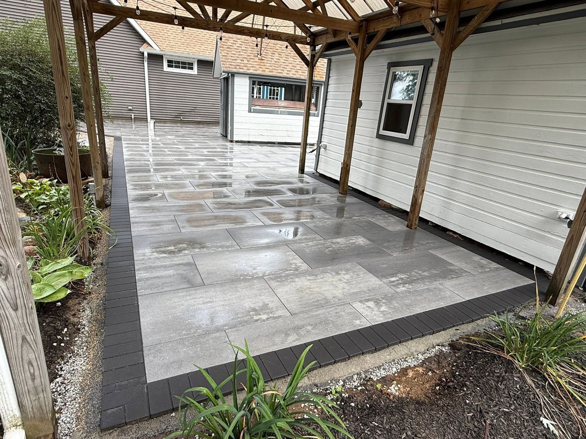 Covered stone patio beside a white house, with dark edging and a wooden roof structure.