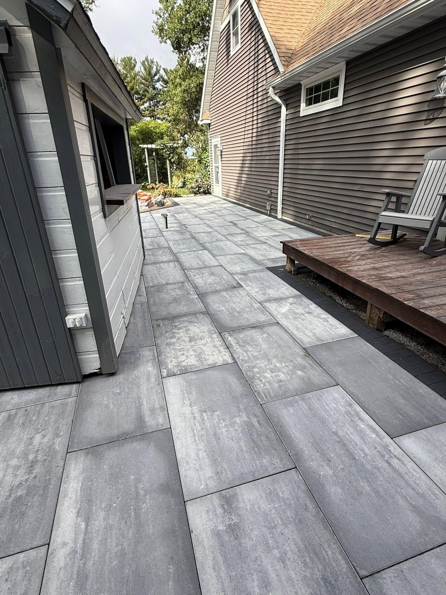 Narrow stone patio walkway between gray house and brown siding, with a small wooden deck and chair.