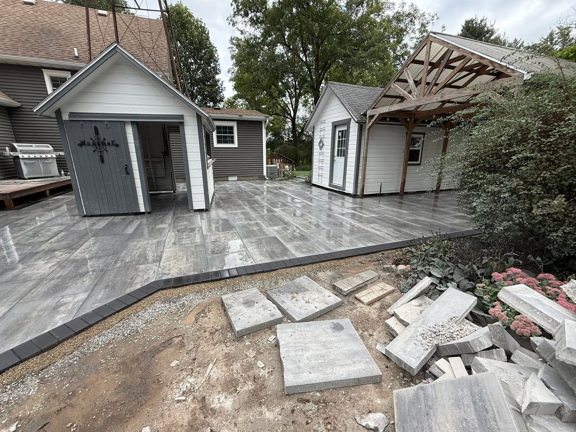 Backyard patio with gray stone pavers, sheds, and a partially dismantled deck area with debris.