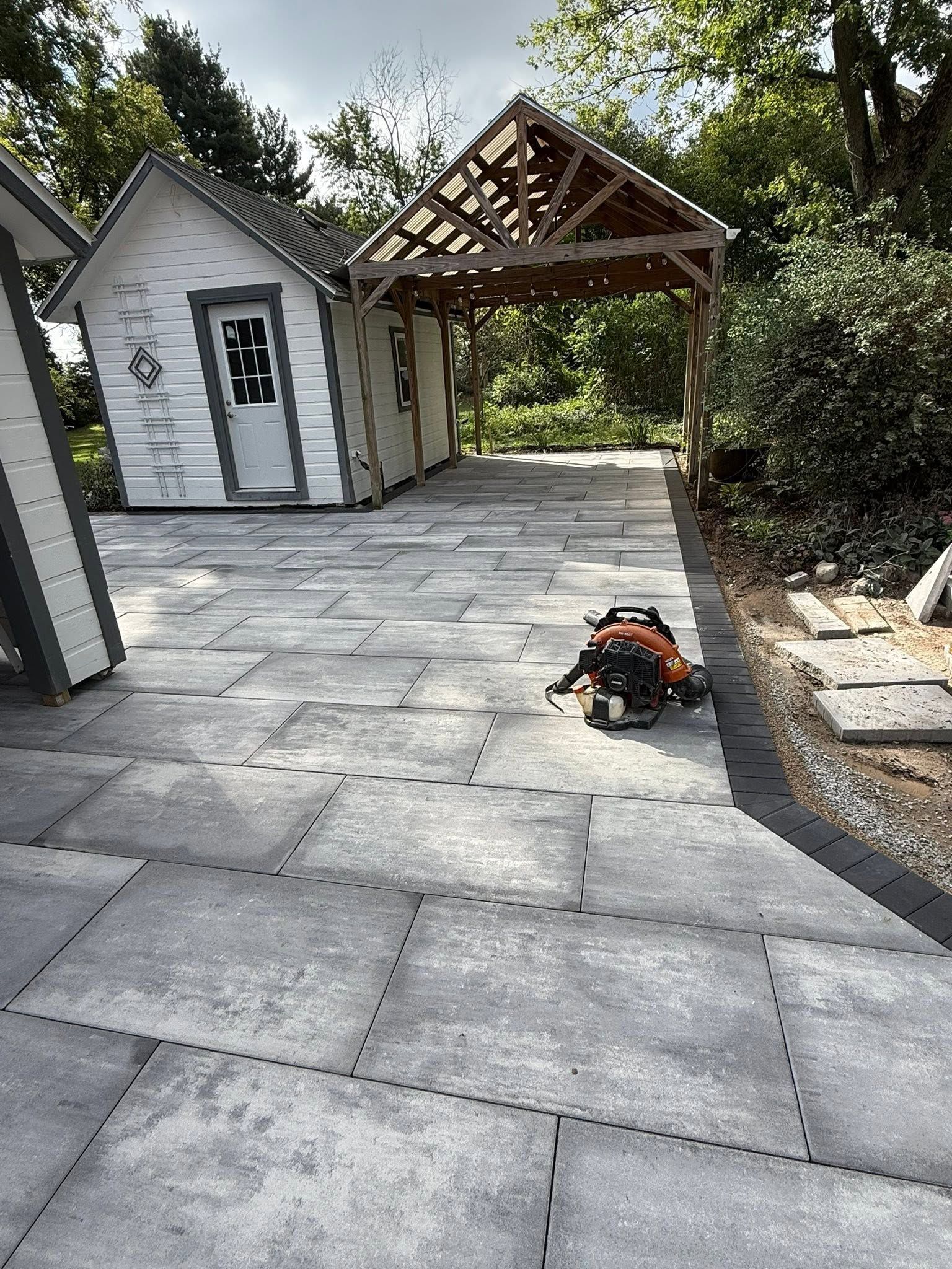 Stone patio leading to a small white shed and pergola, with a tool bag on the ground.